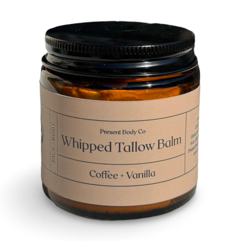 An amber glass jar of face body balm with a black lid and a beige paper label reading 'Present Body Co Whipped Tallow Balm Coffee + Vanilla'.
