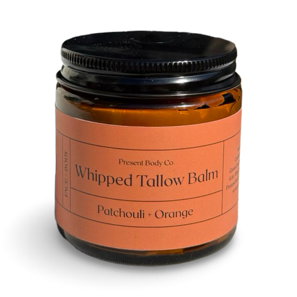 A jar of Present Body Co Whipped Tallow Balm in Patchouli + Orange scent, featuring an amber glass jar with a black lid and a peach-colored label.