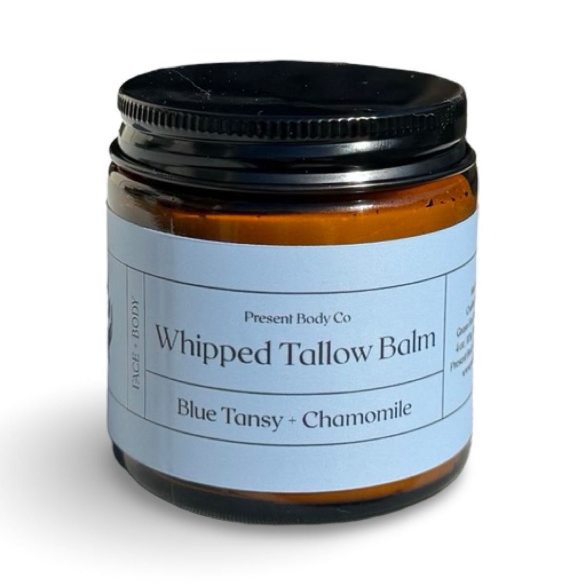 A jar of Present Body Co Whipped Tallow Balm in the Blue Tansy + Chamomile scent, featuring an amber glass jar, a black lid, and a light blue label.