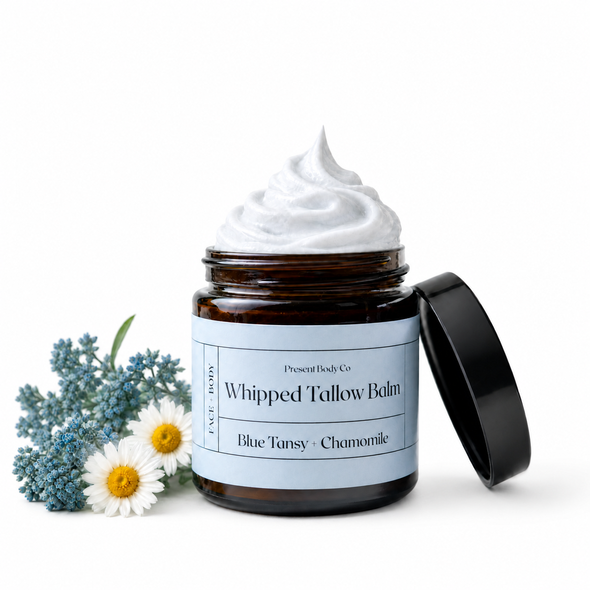 A jar of whipped tallow balm with white cream on top, shown open with its black lid leaning against it on a white background. Blue flowers and white chamomile flowers are placed beside the jar.