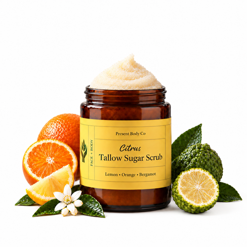A jar of Present Body Co. Citrus Tallow Sugar Scrub with sugar scrub visible at the top, styled with citrus fruits, bergamot, and green leaves on a white background.