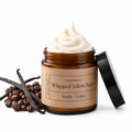 An open amber glass jar of whipped tallow balm with a black lid leaning against it, shown on a white background with vanilla beans and coffee beans beside it. The label reads 'Present Body Co.
