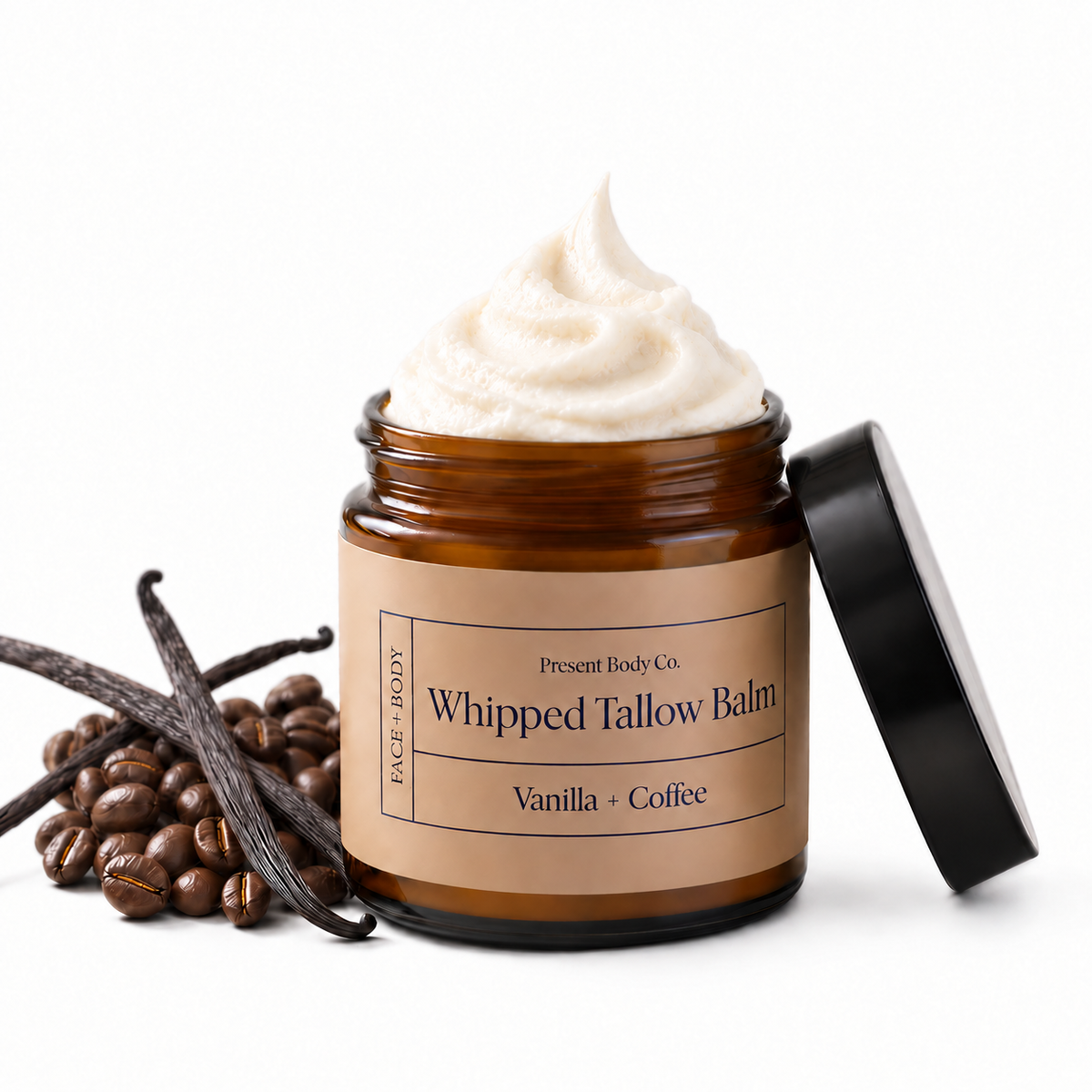 An open amber glass jar of whipped tallow balm with a black lid leaning against it, shown on a white background with vanilla beans and coffee beans beside it. The label reads 'Present Body Co.