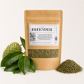 A kraft paper stand-up pouch labeled DEFENDER by PowerHerbs Co, shown on a clean white background with green leaves and a spiky green fruit on the left and a small pile of dried green contents on the right.