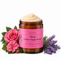 An amber glass jar of Present Body Co. Floral Tallow Sugar Scrub with a pink label, filled with beige sugar scrub and surrounded by fresh pink roses, green leaves, and purple lavender sprigs on a white background.