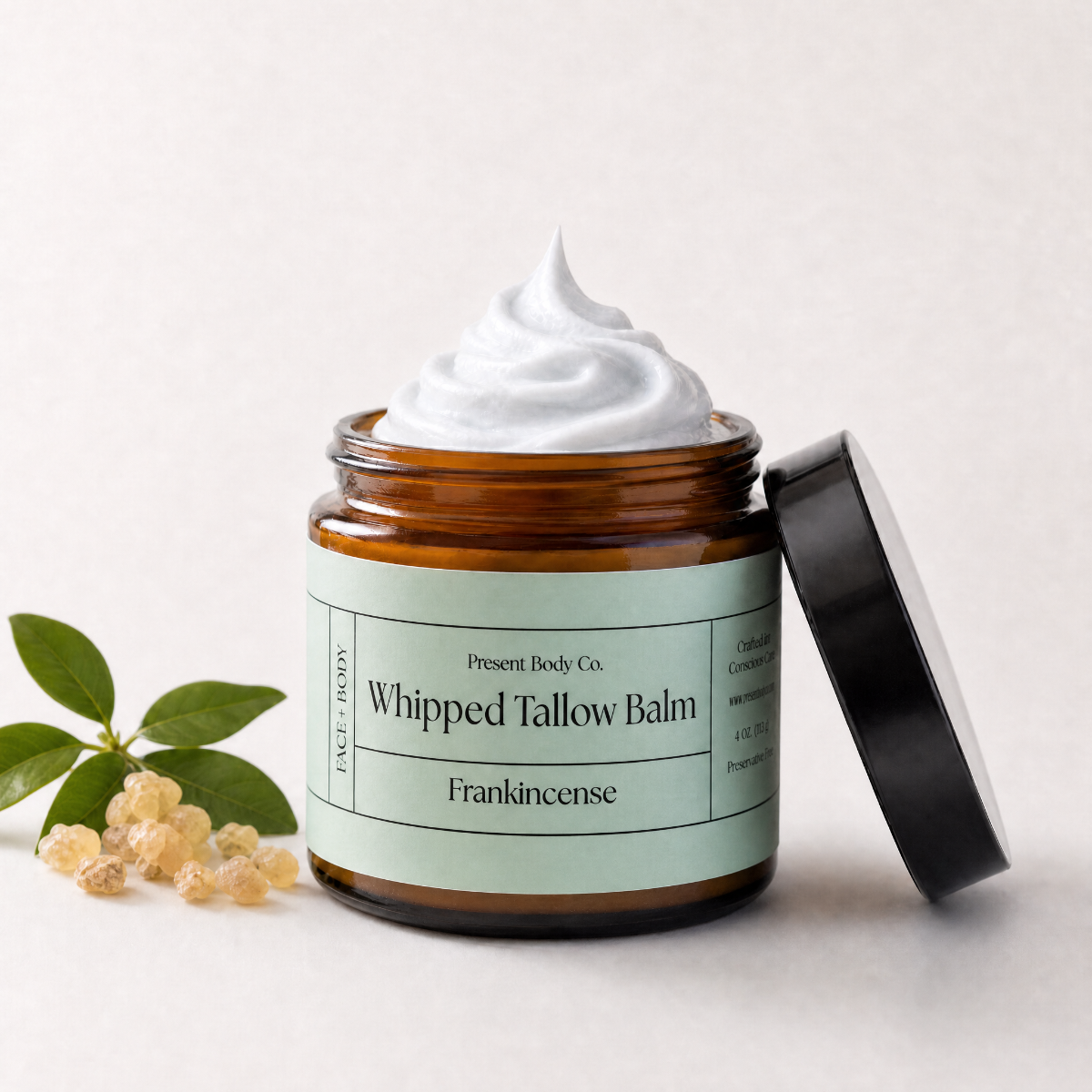 An open amber glass jar of whipped tallow balm with a black lid leaning against it, topped with a swirl of white balm. The label reads 'Present Body Co.
