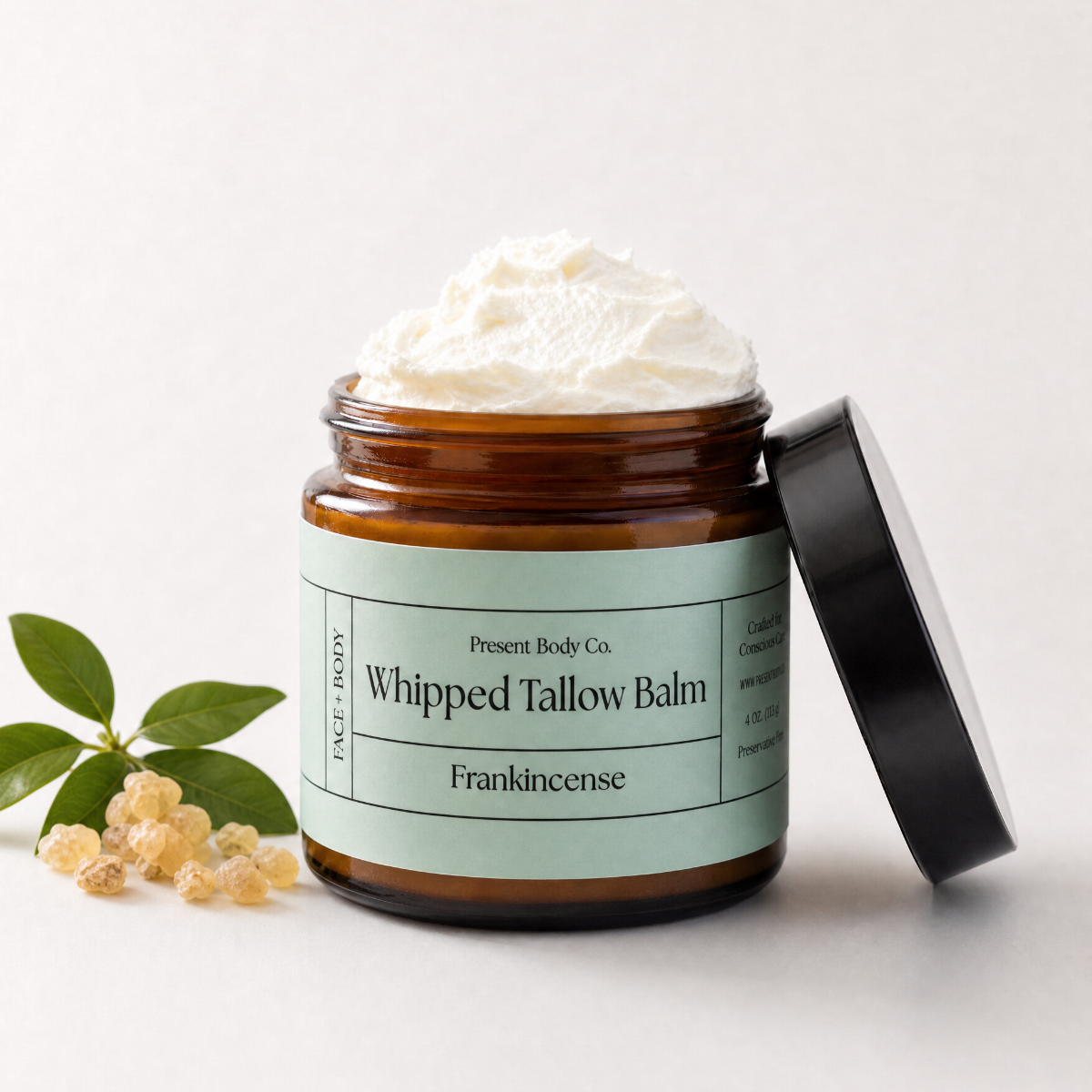 An amber glass jar of whipped tallow balm by Present Body Co. with a pale green label and a black lid resting beside it.