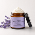 An amber glass jar of lavender whipped tallow balm by Present Body Co. with a black lid leaning against it, topped with white balm, set on a light neutral background with lavender sprigs.