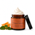 An open amber glass jar of whipped tallow balm with a black lid leaning against it, topped with a swirl of off-white balm. The jar has an orange label reading 'Present Body Co.