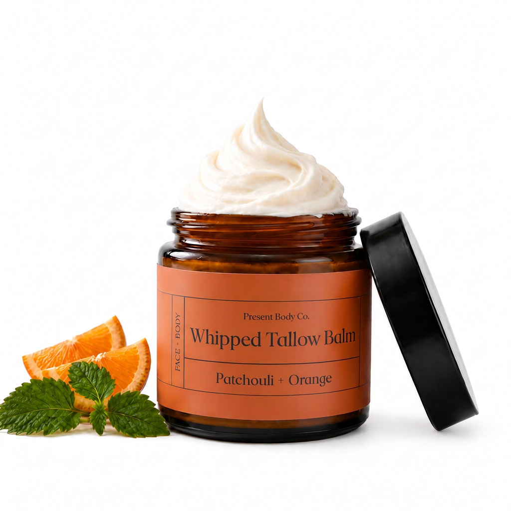 An open amber glass jar of whipped tallow balm with a black lid leaning against it, topped with a swirl of off-white balm. The jar has an orange label reading 'Present Body Co.