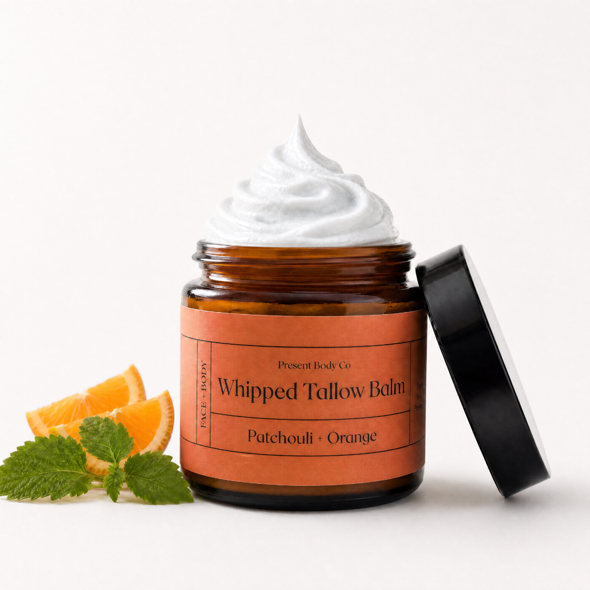 An open amber glass jar of Present Body Co Whipped Tallow Balm with a black lid leaning against it, topped with white balm cream. Slices of orange and mint leaves are placed on the left side.