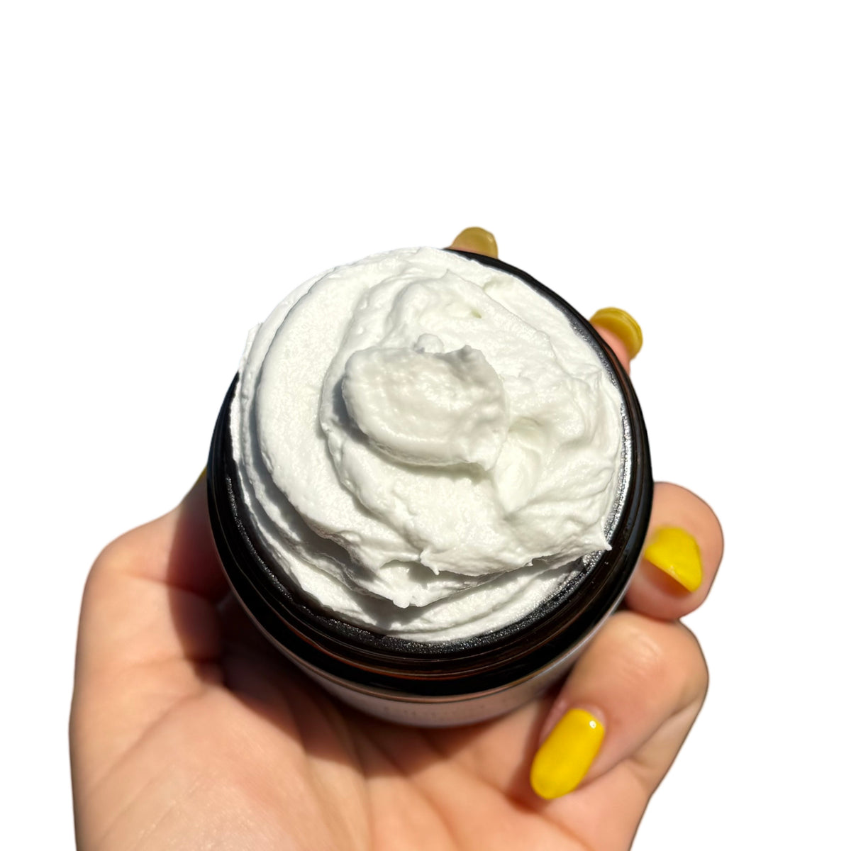 A hand with yellow fingernails holds an open dark amber jar filled with thick white cream against a plain white background.