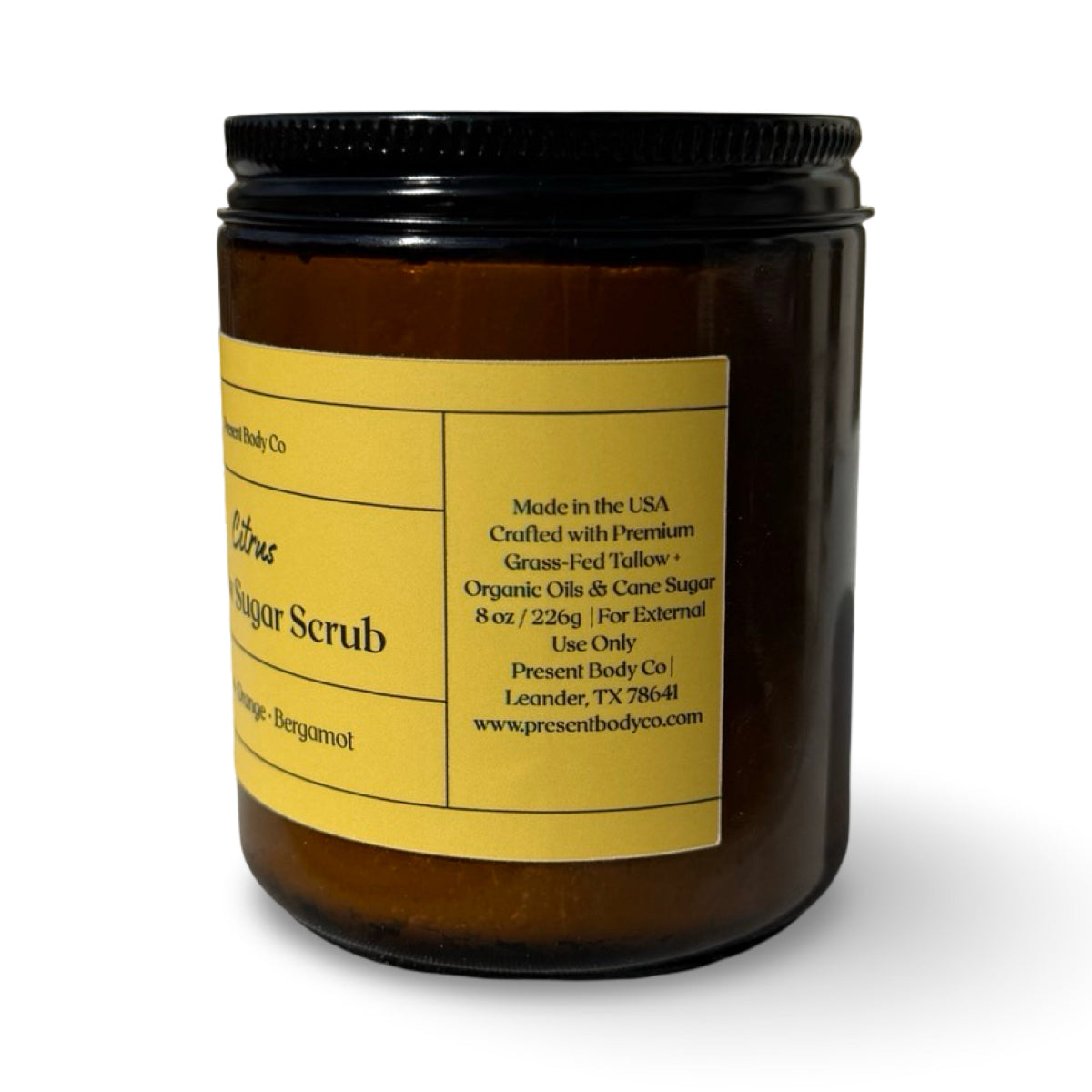 An amber glass jar of Present Body Co. Citrus Tallow Sugar Scrub with a black screw-on lid and a yellow label, shown on a white background.