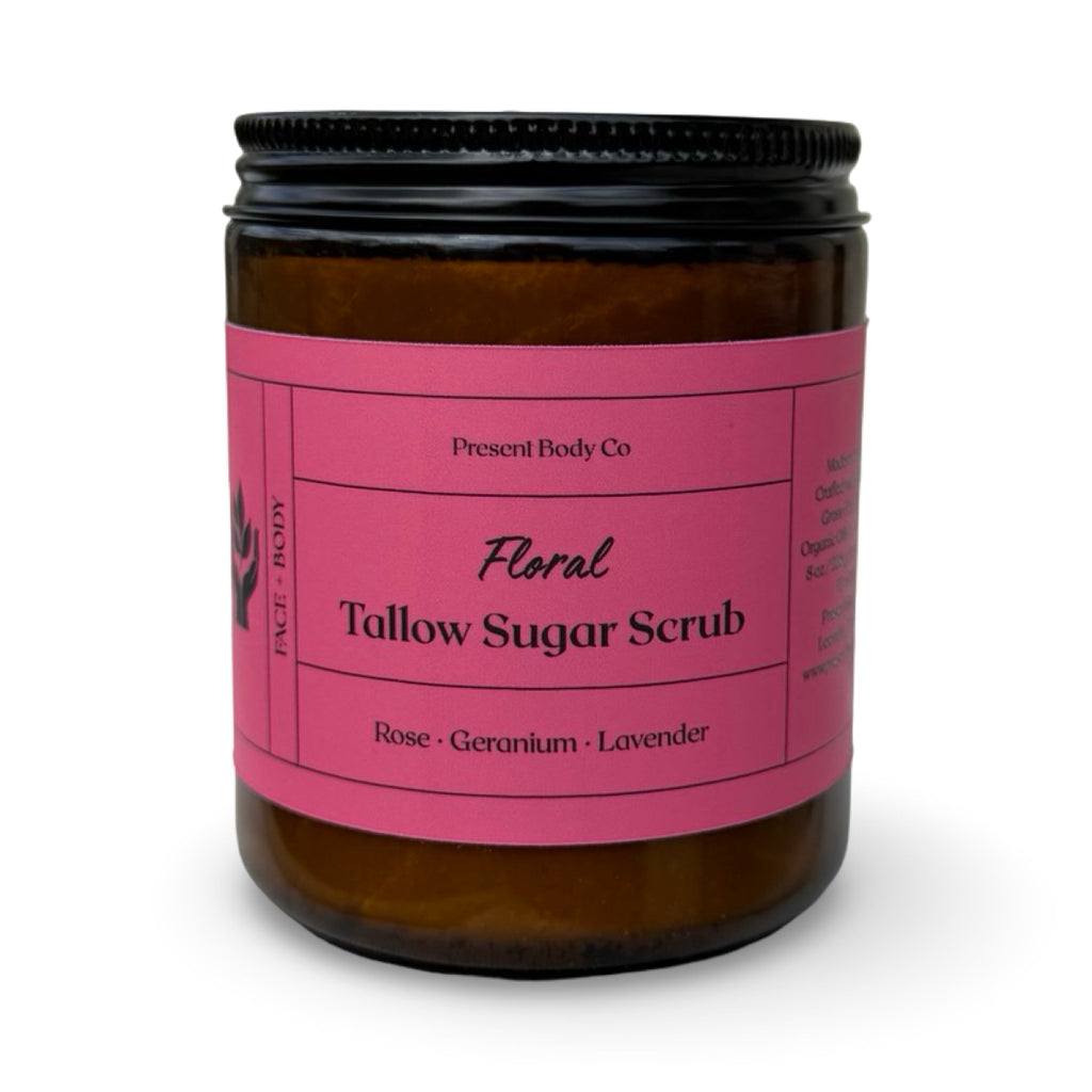 An amber glass jar of Present Body Co. Floral Tallow Sugar Scrub with a bright pink label and a black screw-on lid, set against a plain white background.