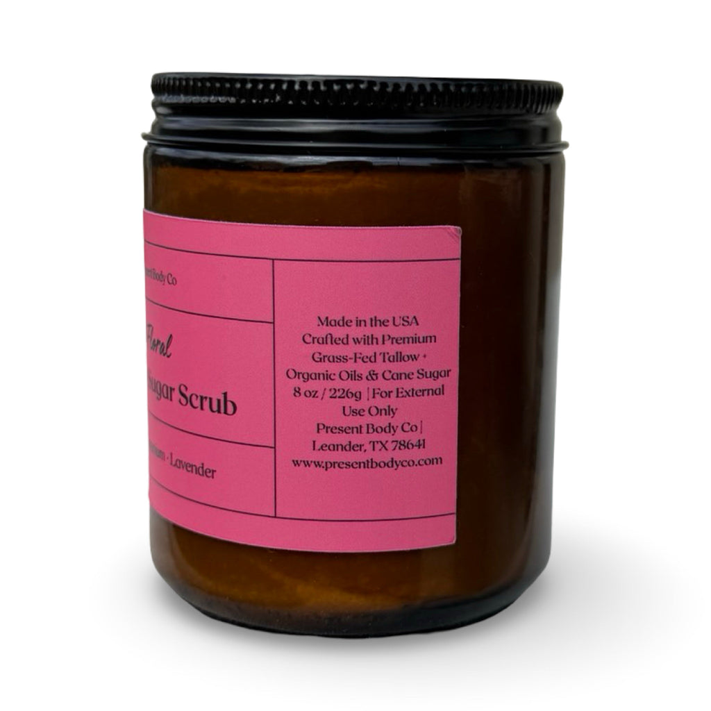 An amber glass jar containing a lavender body sugar scrub, featuring a bright pink label with black text and a black screw-on lid, set against a plain white background.