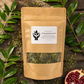 A kraft paper stand-up pouch labeled 'Present Body Co. Pomegranate Leaf' sits on a rustic wooden surface surrounded by green leaves and a sliced pomegranate half.