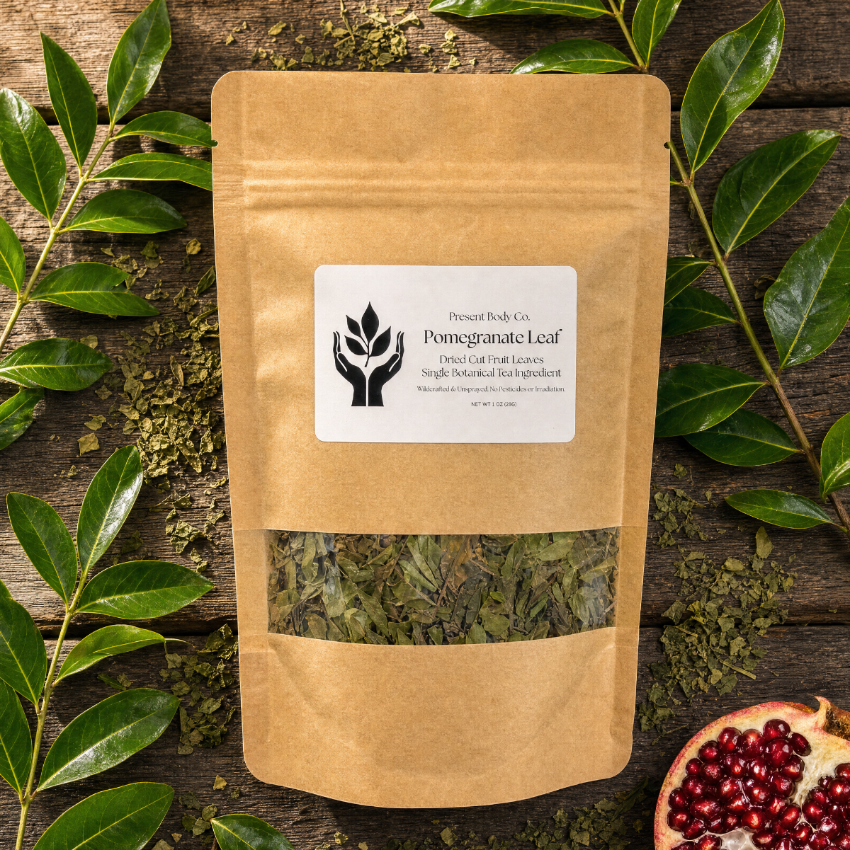 A kraft paper stand-up pouch labeled 'Present Body Co. Pomegranate Leaf' sits on a rustic wooden surface surrounded by green leaves and a sliced pomegranate half.