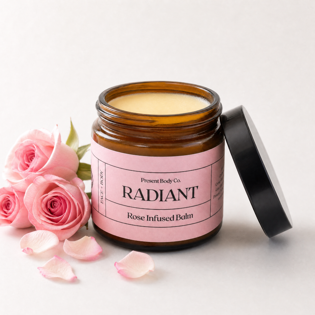 An open amber glass jar of Present Body Co. Radiant Rose Infused Balm with its black lid leaning against it, styled with pink roses and scattered petals on a clean white background.