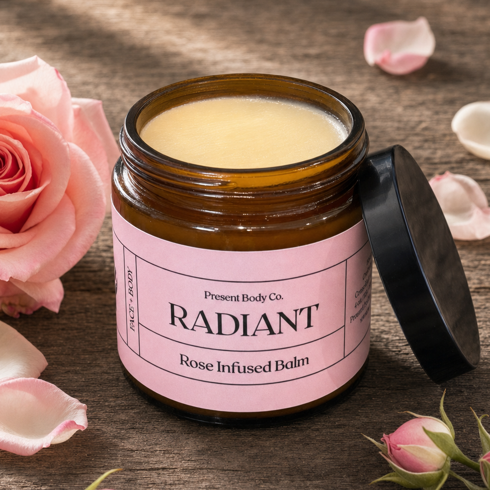 An open amber glass jar of Present Body Co. Radiant Rose Infused Balm with its black lid leaning against it, styled on a rustic wooden surface with pink rose petals and buds.