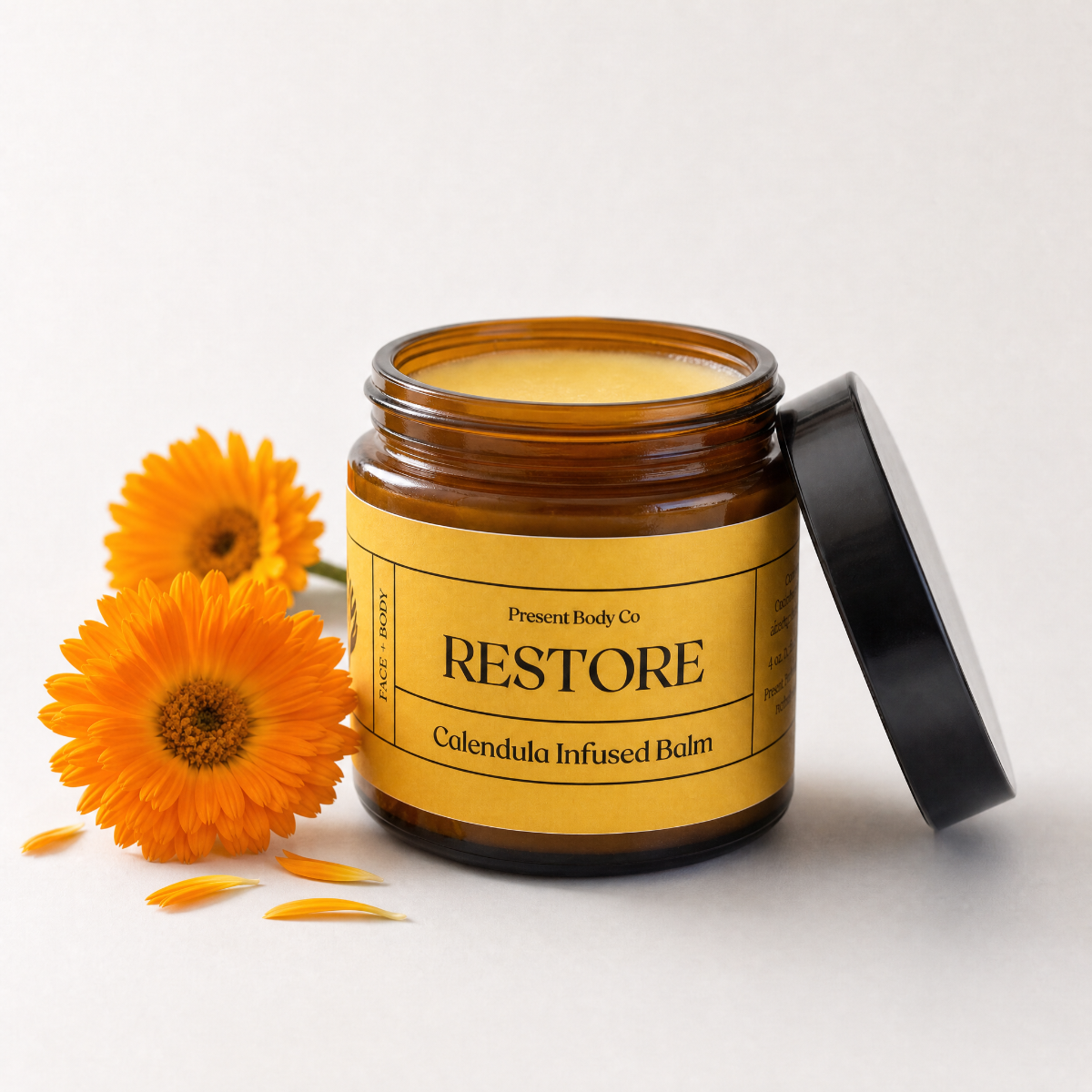An amber glass jar of Present Body Co. Restore Calendula Infused Balm with a yellow label and black lid, styled with two orange calendula flowers on a clean white background.