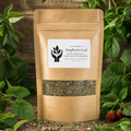 A kraft paper stand-up pouch labeled 'Raspberry Leaf' by PowerBody Co., surrounded by fresh green leaves and a few red raspberries on a rustic wooden surface.