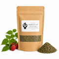 A kraft paper stand-up pouch of Raspberry Leaf by Present Body Co., shown on a white background with green leaves and red raspberries on the left and a small pile of dried green leaves on the right.