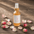 A bottle of Rose Water fragrance by Dreamy Soft Co, filled with amber liquid and topped with a gold cap, set on a rustic wooden surface surrounded by pink rose petals and buds.