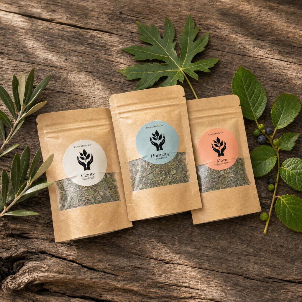 Three stand-up kraft paper pouches of dried herbs arranged on a rustic wooden surface, with green leaves placed around them. The center pouch has a light blue label, the left pouch has a white label, and the right pouch has a peach label, all showing the same botanical logo.