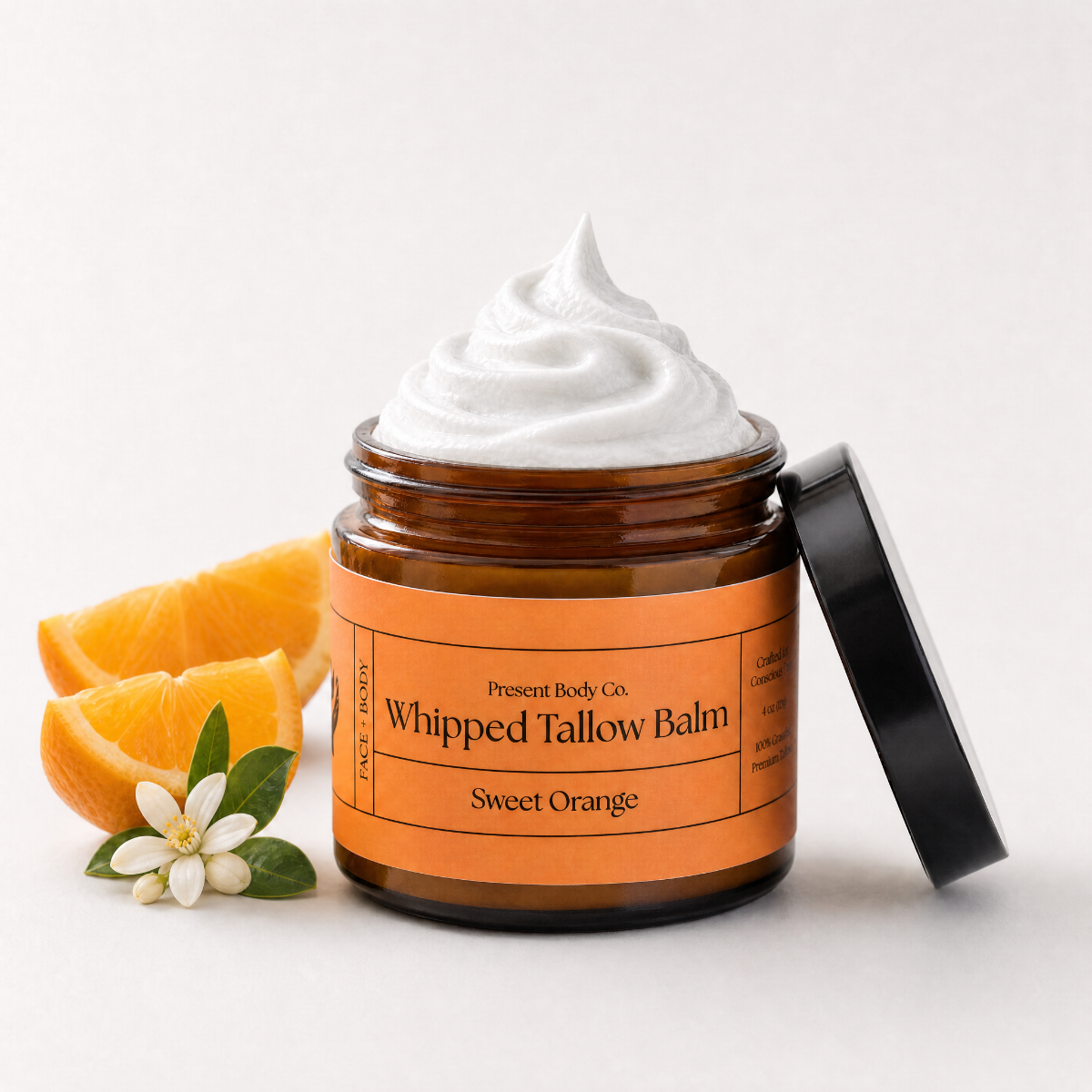 An amber glass jar of whipped tallow balm with a black lid leaning against it, topped with a swirl of white balm. The jar has an orange label reading 'Present Body Co.