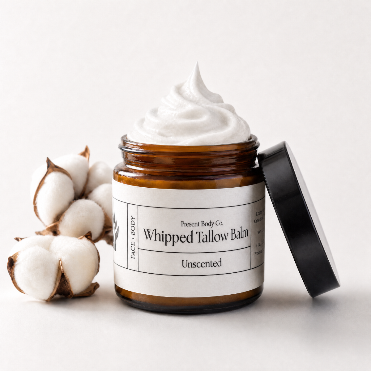 An open amber glass jar of whipped tallow balm with a black lid leaning beside it, topped with a swirl of white balm. Cotton boll props sit on the left, and the label reads Present Body Co.