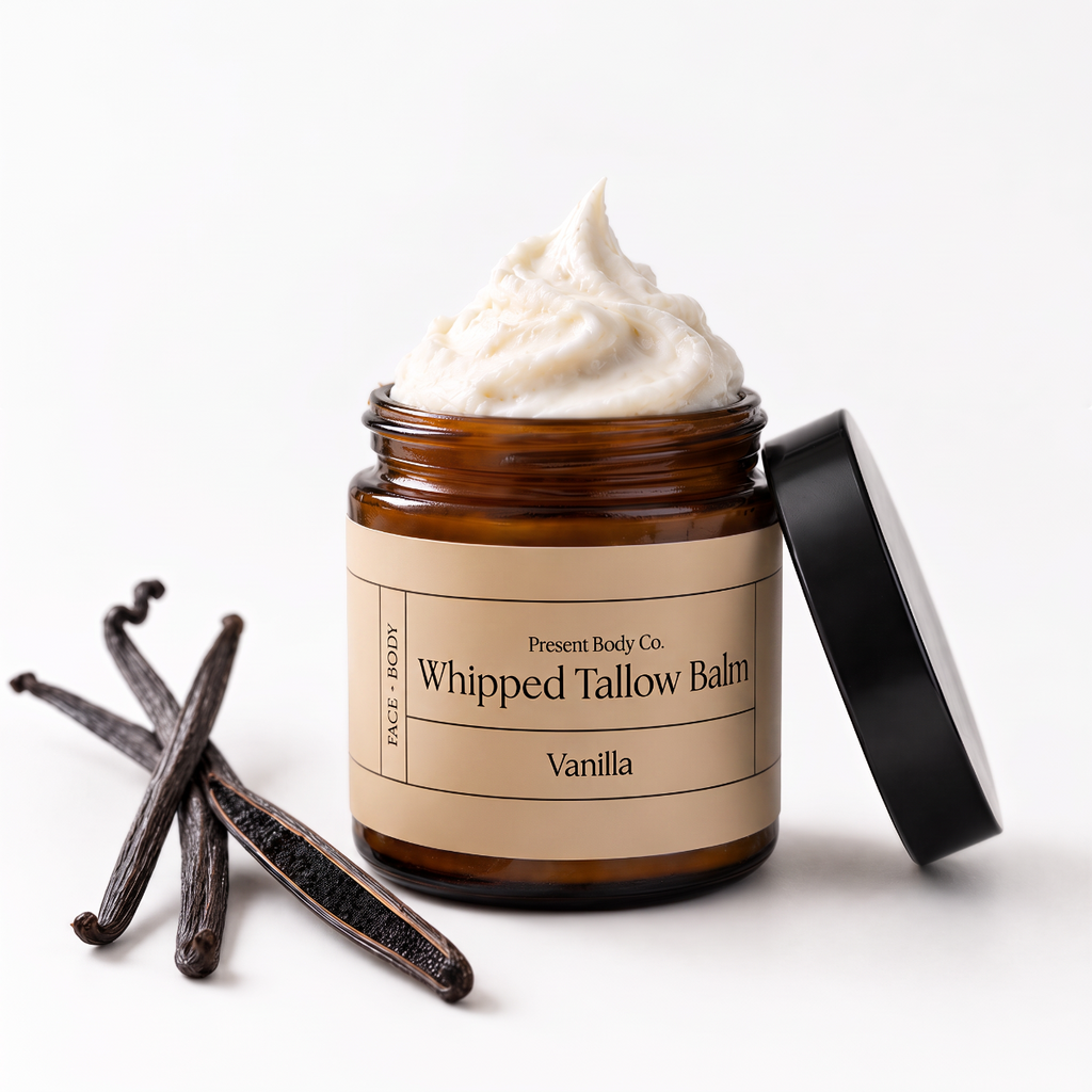 An open amber glass jar of whipped body balm with a black lid leaning against it, shown on a clean white background with vanilla beans beside the product. The label reads 'Present Body Co.