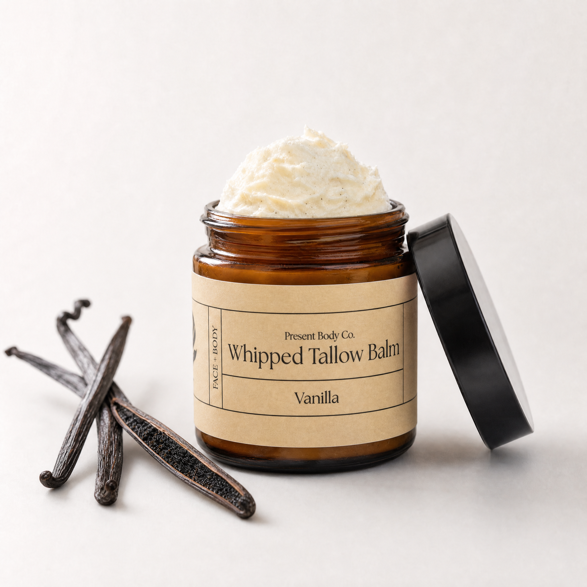 An amber glass jar of whipped tallow body balm with its black lid leaning against it, shown open with cream-colored balm inside. Vanilla beans are placed beside the jar on a light neutral background.