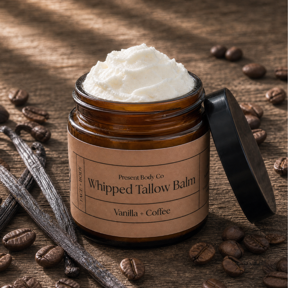 An amber glass jar of Present Body Co. Whipped Tallow Balm in Vanilla + Coffee scent, filled with white whipped balm and shown with its black lid leaning against it on a rustic wooden surface scattered with coffee beans and vanilla beans.