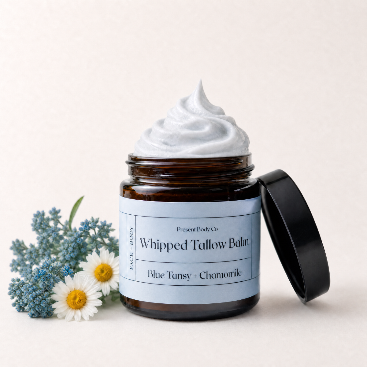 An amber glass jar of Present Body Co Whipped Tallow Balm with a light blue label and black lid, topped with white balm, styled with blue flowers and white daisies on a light beige background.